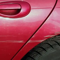 Kolizja na parkingu podczas cofania: głębokie rysy na czerwonym lakierze, od klamki po oponę. Mandat za nieostrożność.