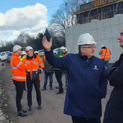 Grupa mężczyzn w kaskach i kamizelkach odblaskowych ogląda postęp prac przy budowie obwodnicy Krakowa. W tle koparka i betonowa konstrukcja.