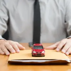 Czerwony samochodzik na kopercie, obok dłonie mężczyzny w koszuli i krawacie. Dokumenty w kopercie mogą być kartą pojazdu.