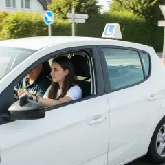 Młoda dziewczyna z instruktorem za kierownicą białego samochodu z literą L, ucząca się jeździć. W Polsce prawo jazdy można uzyskać od 18 lat.