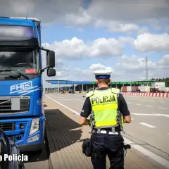 Policjant w kamizelce odblaskowej przy niebieskiej ciężarówce Volvo FH13. W tle przejście graniczne, kategoria c.