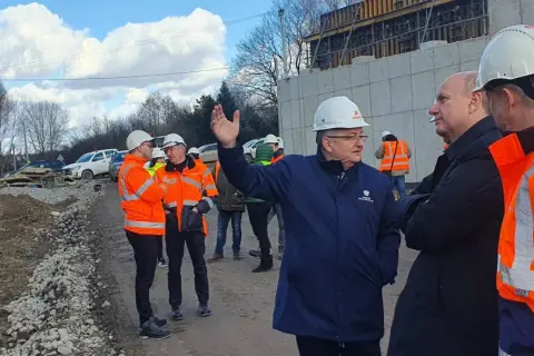 Grupa mężczyzn w kaskach i kamizelkach odblaskowych ogląda postęp prac przy budowie obwodnicy Krakowa. W tle koparka i betonowa konstrukcja.