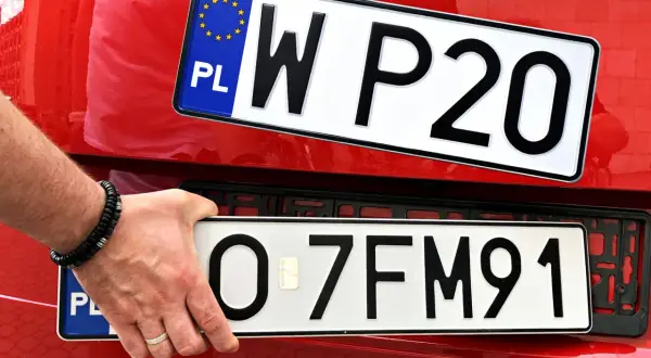 Ręka z bransoletką montuje tablicę rejestracyjną z napisem "O 7FM91" na czerwonym samochodzie. Obok widoczna jest inna tablica "W P20".