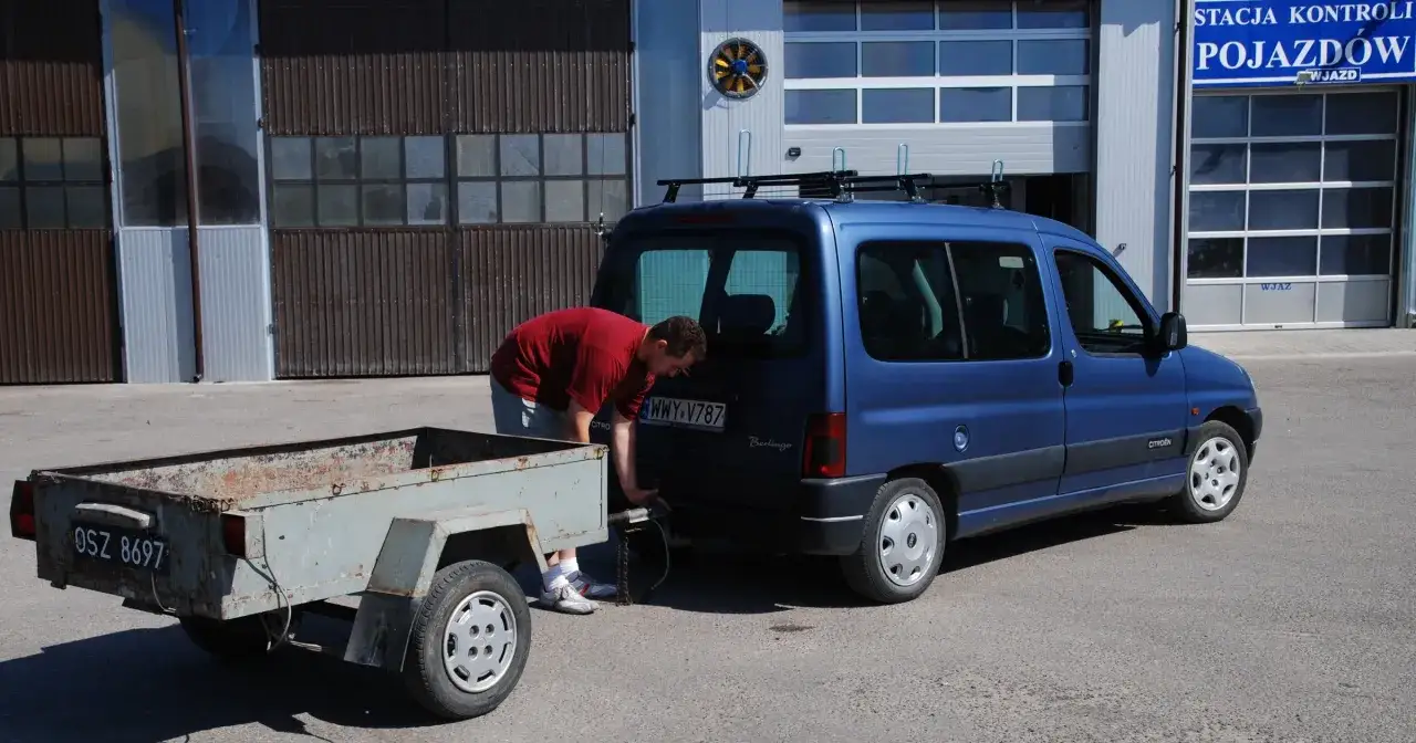 Mężczyzna podłącza starą przyczepę do niebieskiego Citroena Berlingo. Taka przyczepa na kat B jest idealna do przewozu drobnych ładunków.