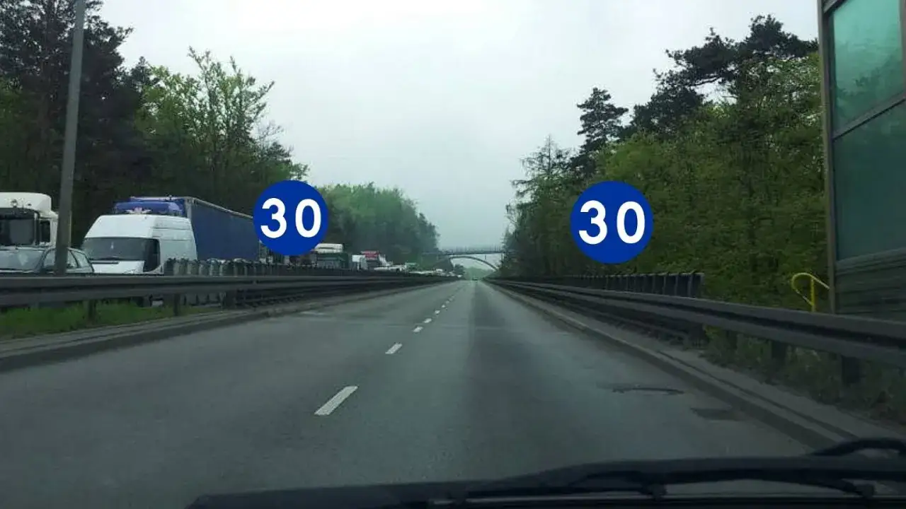 Na autostradzie widać znaki informujące o minimalnej prędkości 30 km/h. Ruch jest spowolniony, stoją ciężarówki.