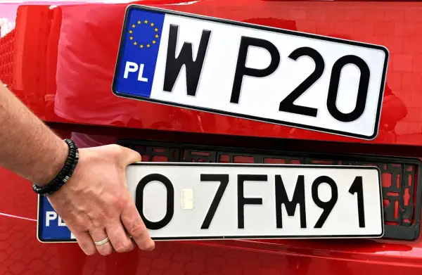 Ręka z bransoletką montuje tablicę rejestracyjną z napisem "O 7FM91" na czerwonym samochodzie. Obok widoczna jest inna tablica "W P20".