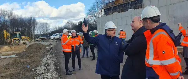 Grupa mężczyzn w kaskach i kamizelkach odblaskowych ogląda postęp prac przy budowie obwodnicy Krakowa. W tle koparka i betonowa konstrukcja.