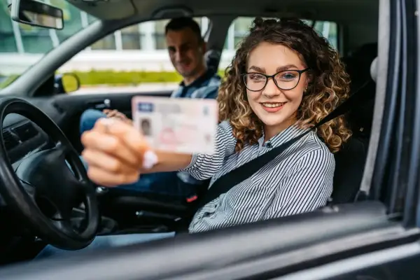 Uśmiechnięta młoda kobieta w okularach pokazuje prawo jazdy, rozpoczynając swój okres próbny. Za nią siedzi instruktor.